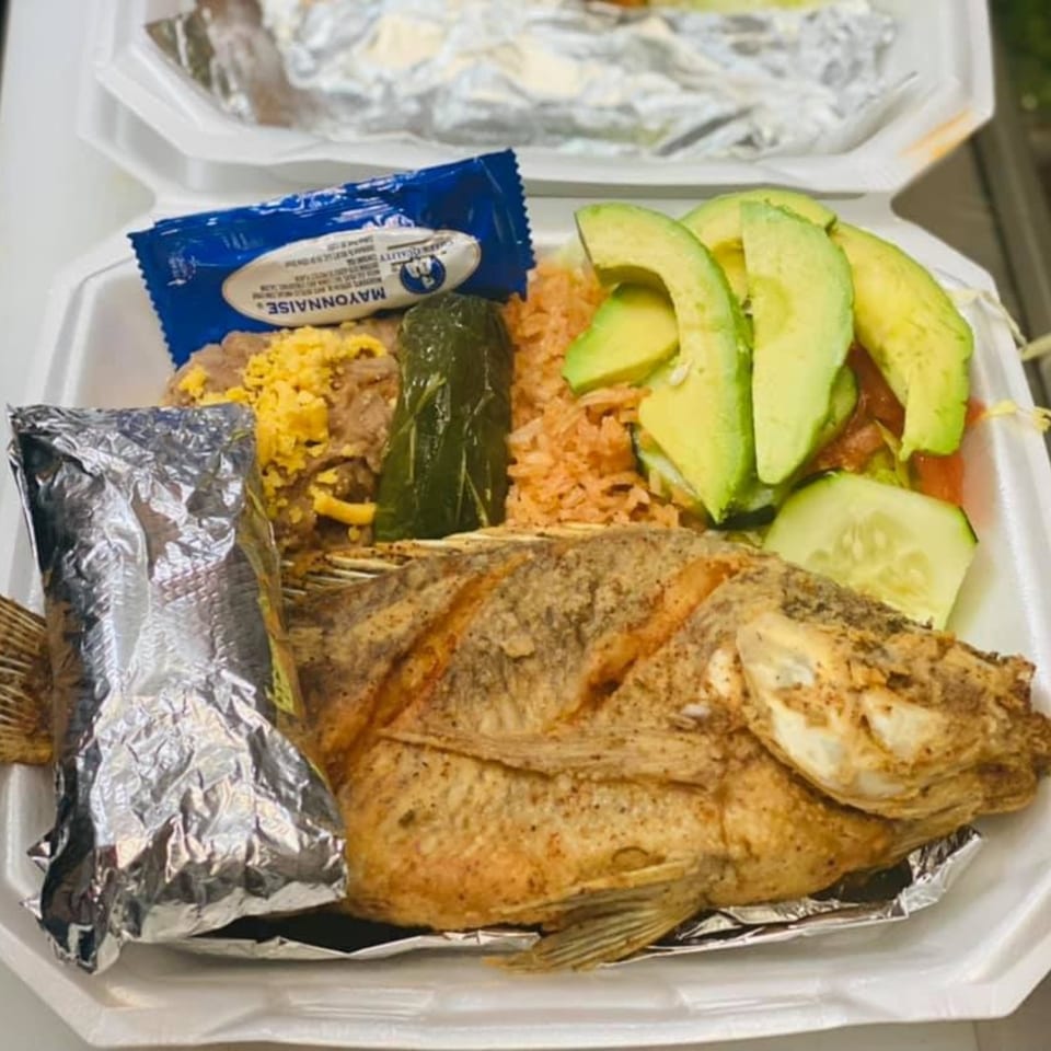 Fried whole fish with rice, beans, avocado, cucumber, and a mayonnaise packet in a takeout container.